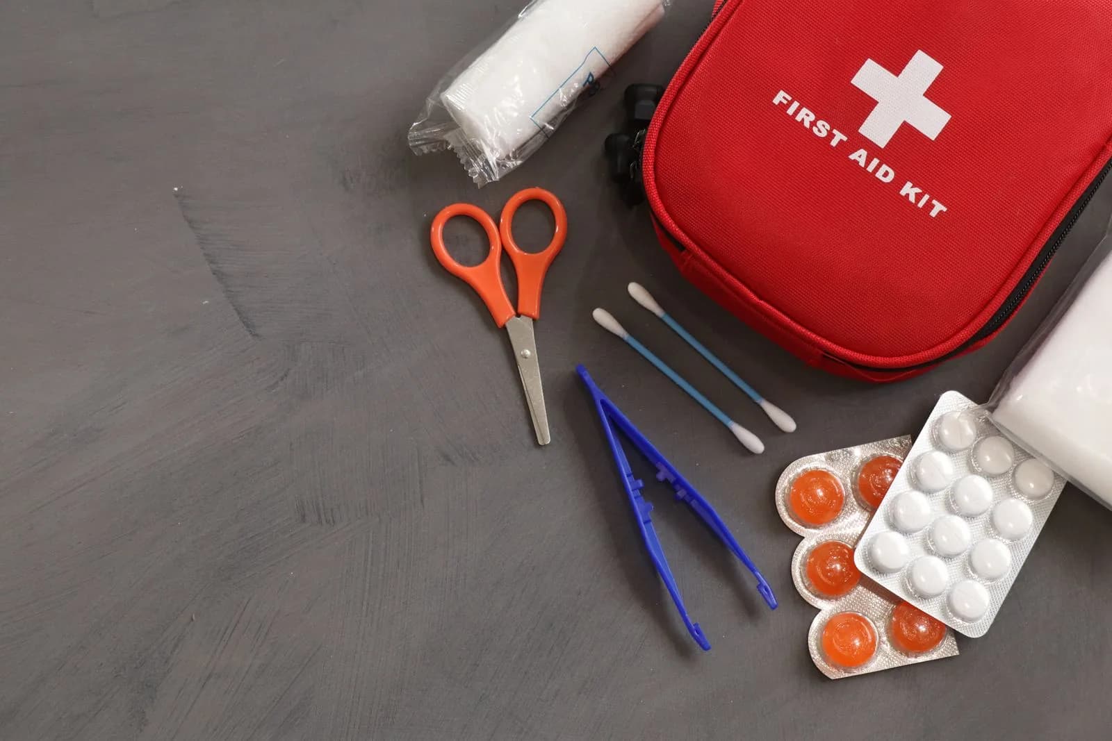 An open first-aid kit with bandages, scissors and emergency supplies arranged on a grey surface.