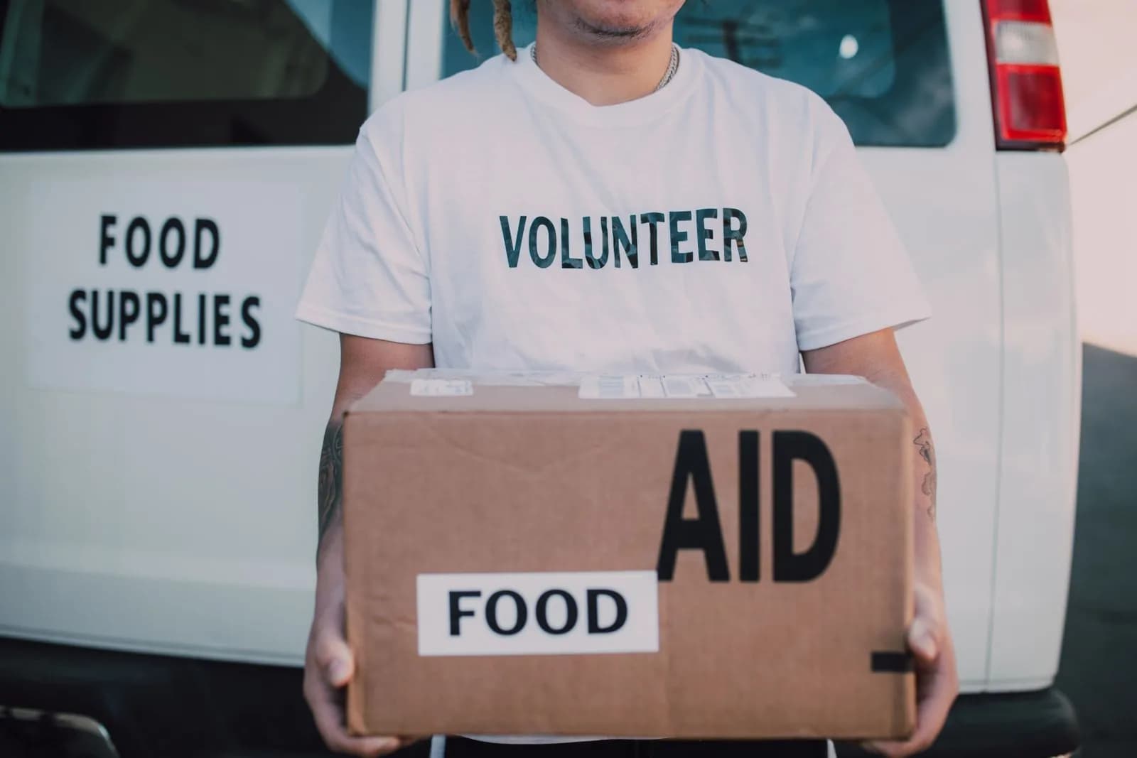 A volunteer holding a box of food and essential supplies beside a vehicle.
